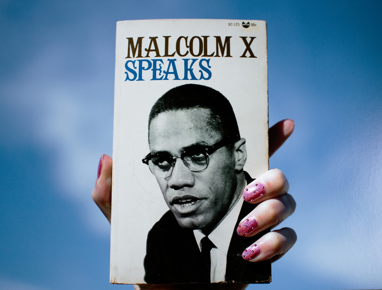 Hand holding up book of 'Malcolm X Speaks' with pink nailpolish against blue backdrop