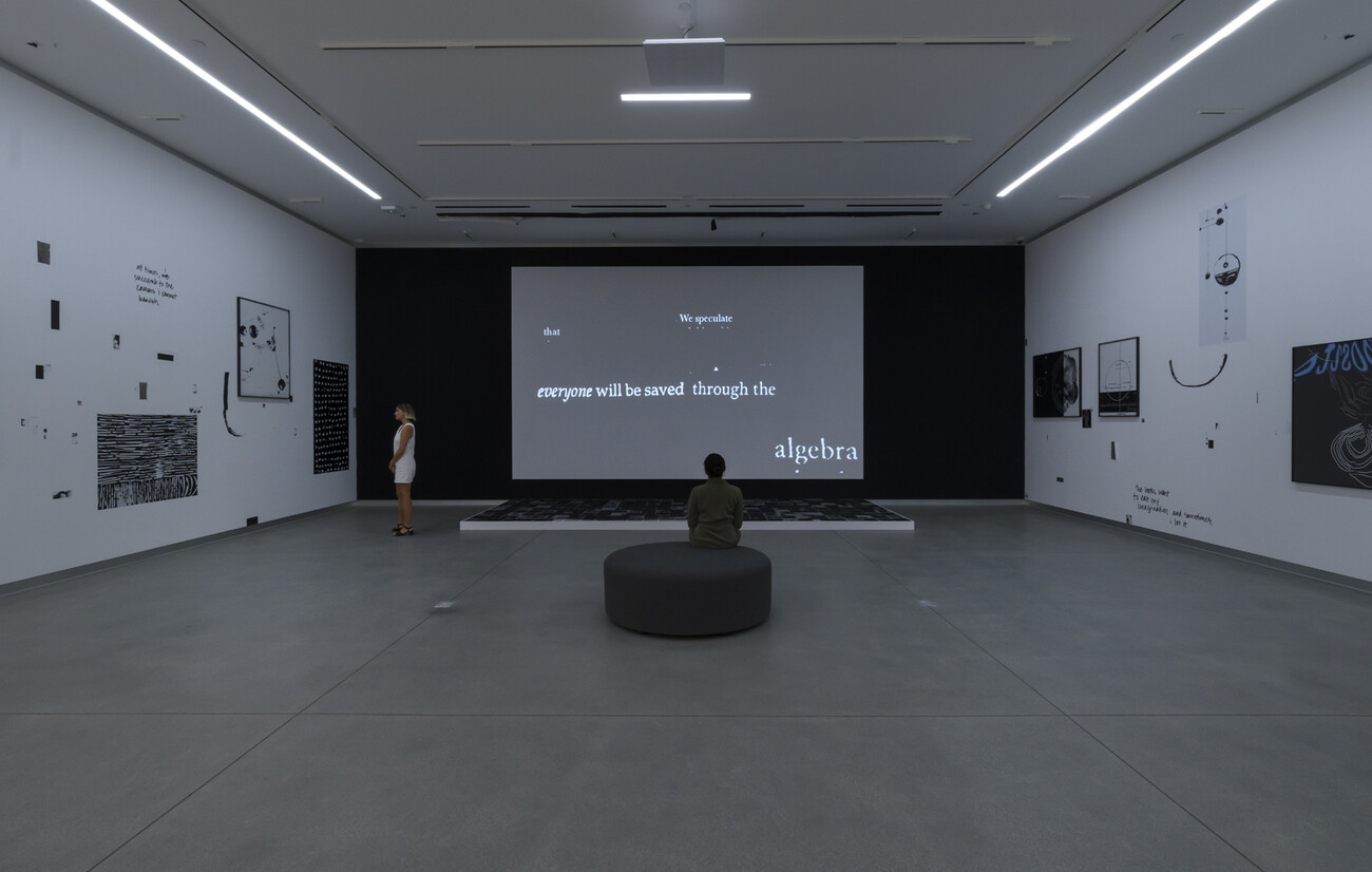 dark room with black and white works and video. two people sand and sit in the room