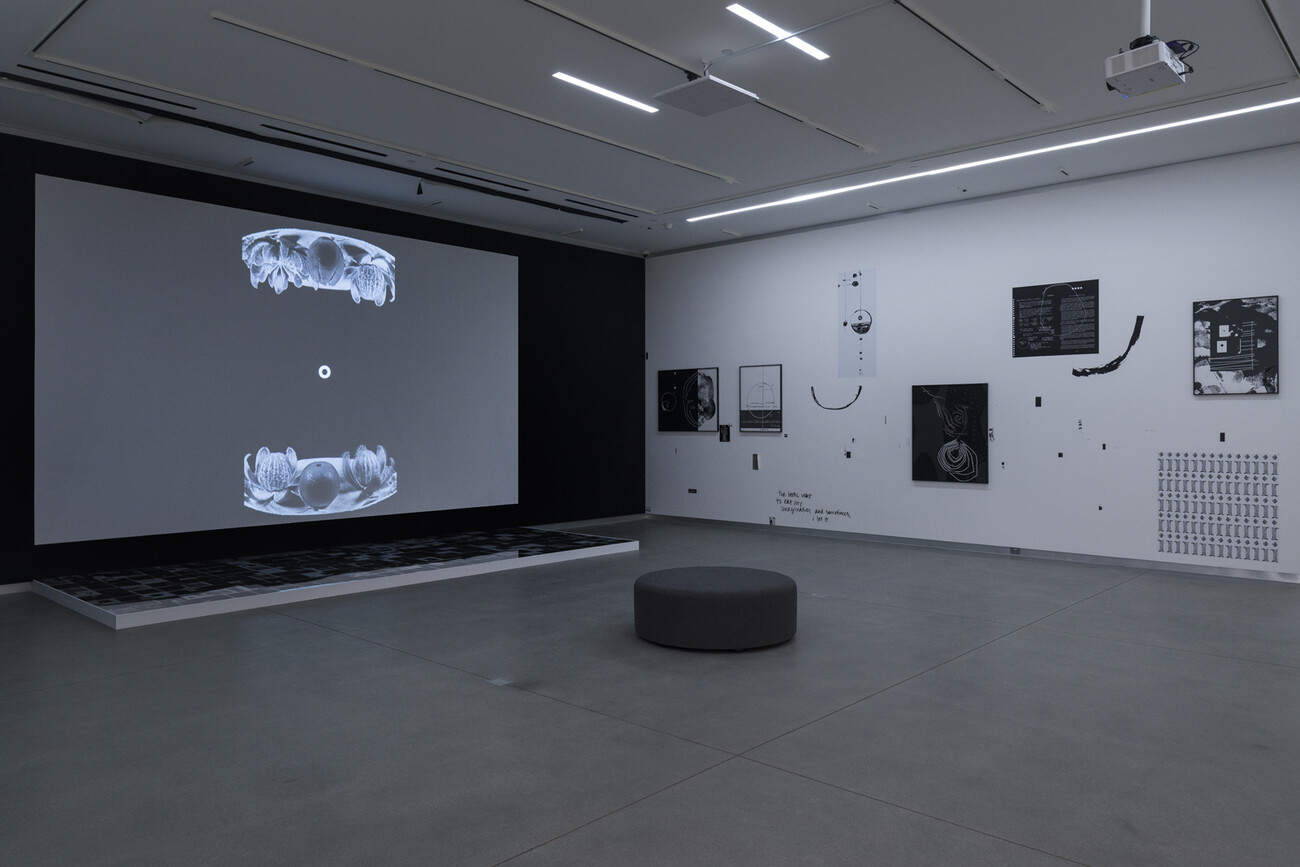 room with black and white images randomly on wall. Video playing on front wall. Round pouf in the center of the room