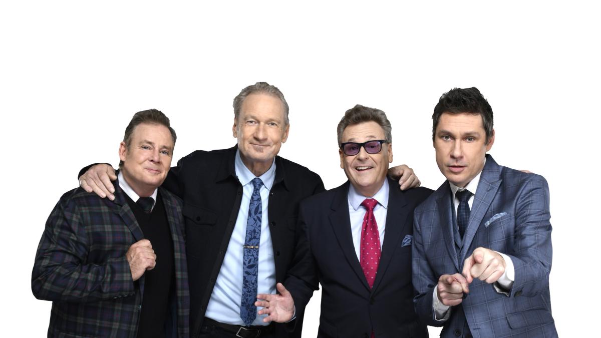The four all-male cast of Whose Line Is It Anyway, dressed in suits, pose on a white background facing the camera.
