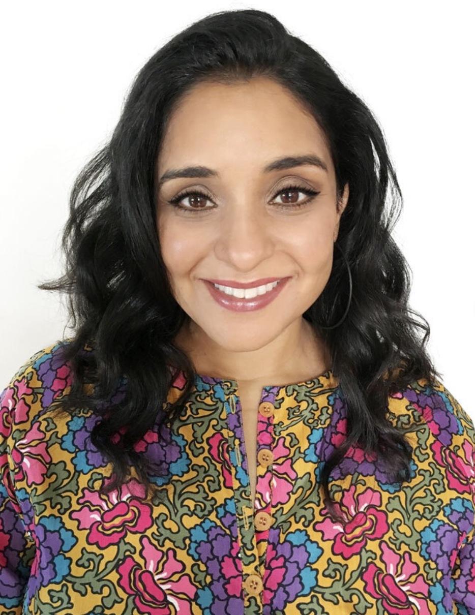 Photo of creative writing workshop leader, local author Neela Banerjee. An Indian-American woman with long, wavy black hair and a bright yellow, purple, blue, pink, and green floral top smiles at the camera.