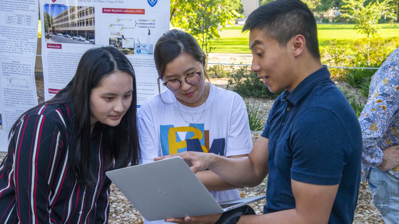 Photo of a student holding an open laptop showing the screen to two additional students with a research poster displayed behind them.